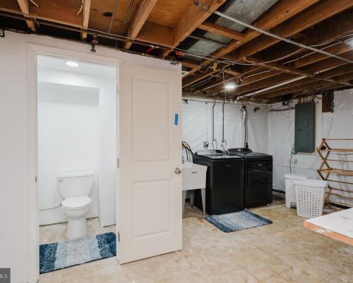 Basement utility area with a washer and dryer, utility sink, laundry basket, exposed ceiling joists, and a small bathroom with a toilet nearby.