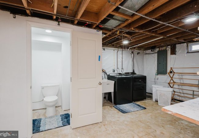 Basement utility area with a washer and dryer, utility sink, laundry basket, exposed ceiling joists, and a small bathroom with a toilet nearby.