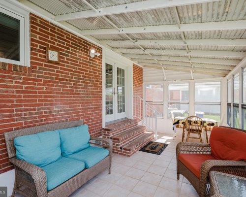Sunroom with brick wall, teal cushions on a wicker sofa, red-cushioned chair, and a glass-top table by a row of windows.