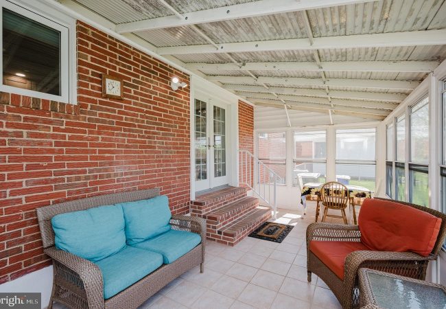 Sunroom with brick wall, teal cushions on a wicker sofa, red-cushioned chair, and a glass-top table by a row of windows.