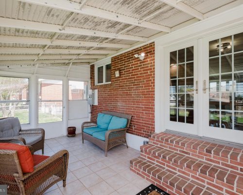 Brick-walled screened porch with wicker seating and turquoise cushions, brick steps to glass French doors on a sunny day behind white frames.