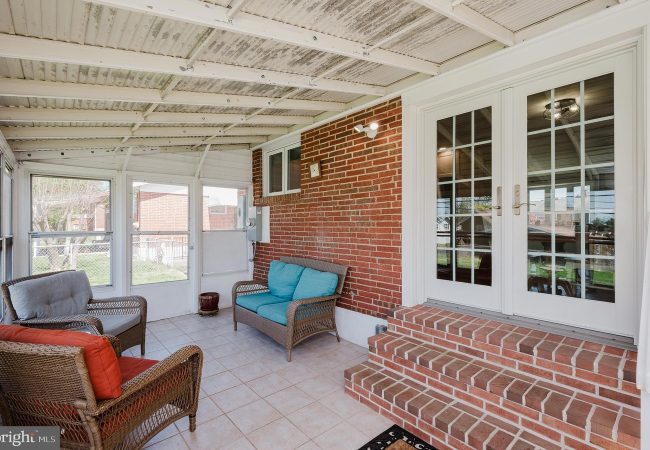 Brick-walled screened porch with wicker seating and turquoise cushions, brick steps to glass French doors on a sunny day behind white frames.