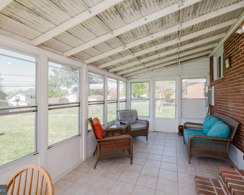Sunroom or enclosed porch with wicker chairs, turquoise cushions, and a brick wall, facing a grassy backyard through large windows.