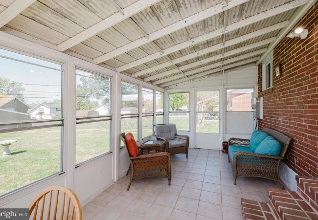 Sunroom or enclosed porch with wicker chairs, turquoise cushions, and a brick wall, facing a grassy backyard through large windows.