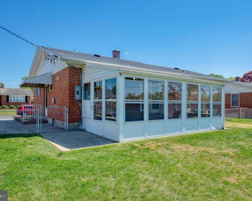 Brick, single-story building with a white-framed glass-enclosed sunroom along the side, set in a grassy yard under a clear blue sky.