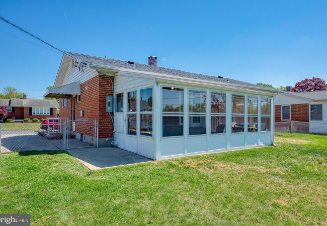 Brick, single-story building with a white-framed glass-enclosed sunroom along the side, set in a grassy yard under a clear blue sky.