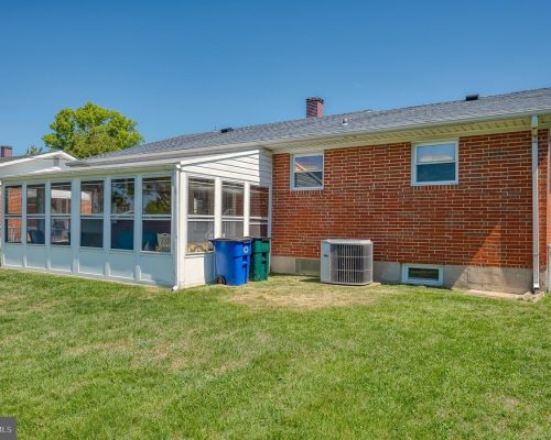 Backyard of a brick house with a white screened porch and blue/green trash bins on the grass in bright daylight.