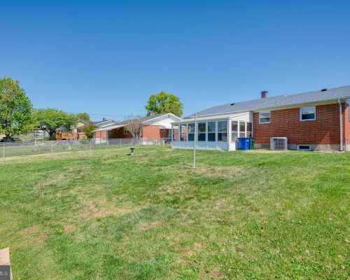 Backyard of a brick house featuring a glass-enclosed sunroom, a green lawn, and a clear blue sky; chain-link fence and trees in the yard.