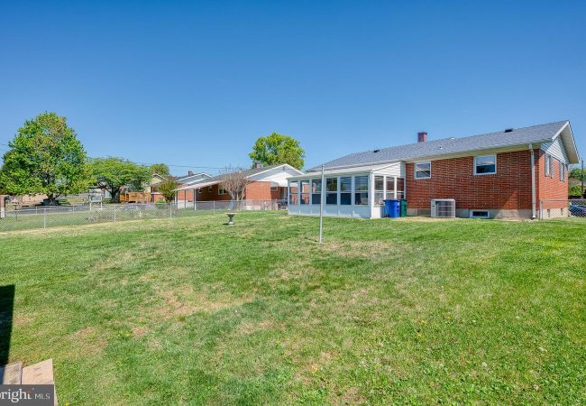 Backyard of a brick house featuring a glass-enclosed sunroom, a green lawn, and a clear blue sky; chain-link fence and trees in the yard.