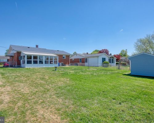 Backyard scene with a brick house featuring a white sunroom, a fenced lawn, and a gray shed on the right under a clear blue sky.