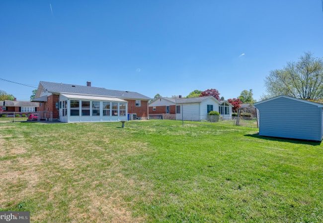 Backyard scene with a brick house featuring a white sunroom, a fenced lawn, and a gray shed on the right under a clear blue sky.