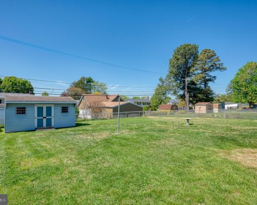 Backyard with a blue shed, clothesline pole, and chain-link fence, green grass under a clear blue sky.