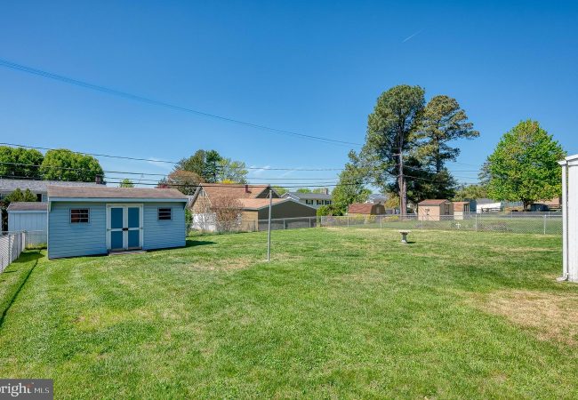 Backyard with a blue shed, clothesline pole, and chain-link fence, green grass under a clear blue sky.
