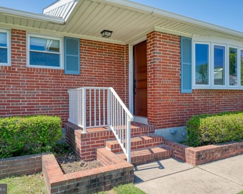 Brick ranch-style home with a white railing and brick steps leading to a wooden front door; blue shutters frame the windows on the brick facade, with trimmed hedges along the walkway.