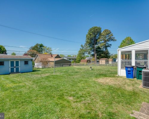 Backyard scene: a small blue shed on the left, a sunroom with several trash bins on the right, a fence, and tall trees in the distance under a clear blue sky.