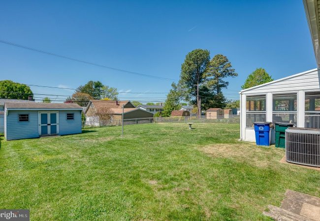Backyard scene: a small blue shed on the left, a sunroom with several trash bins on the right, a fence, and tall trees in the distance under a clear blue sky.