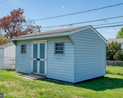 Small blue wooden shed with double doors in a grassy yard, chain-link fence behind, and overhead power lines in a suburban setting.