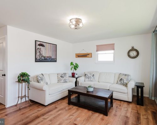L-shaped white sofa set with patterned cushions, a dark wood coffee table, and potted plants in a bright living room.