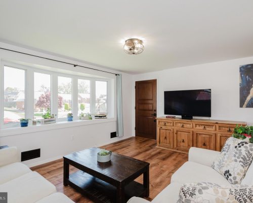 Bright living room with a large bay window, white sofa and spacious wooden coffee table facing a TV on a wooden sideboard.