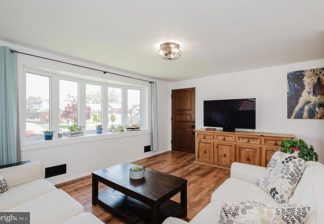Bright living room with a large bay window, white sofa and spacious wooden coffee table facing a TV on a wooden sideboard.